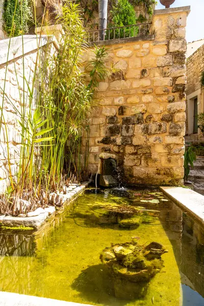 Scicli, Sizilien - İtalya - 04-05-2025: Scicli 'de berrak bir gölet, küçük bir şelale, yeşil bitkiler ve Akdeniz' in altında kırsal taştan bir duvar bulunan parlak bir avlu manzarası.