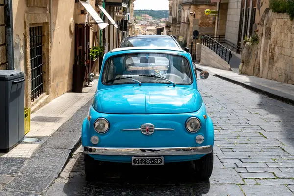 Noto, Sizilien - İtalya - 04-06-2025: Noto 'da bir kaldırım taşı caddesinde park etmiş parlak mavi bir vintage Fiat 500, taş binalar ve eğimli kaldırımlarla çevrili..