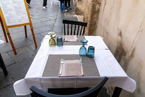 Noto, Sizilien - İtalya - 04-06-2025: Noto 'da iki kişilik açık restoran masası, renkli bardaklar ve çatal bıçaklar desenli örtülerin üzerine beyaz bir masa örtüsü yerleştirilmiş..
