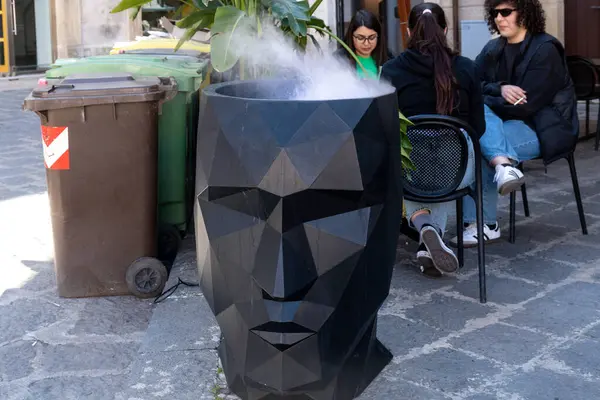 Syrakus, Sizilien - İtalya - 04-08-2025: Geometrik siyah heykel, tepeden yükselen sisi andırıyor, Syracuse caddesinde insanların oturduğu çöp bidonlarına yakın bir yere yerleştirilmiş..