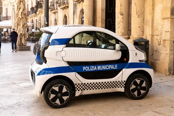 Syrakus, Sizilien - İtalya - 04-08-2025: Syracuse 'un tarihi merkezine park edilmiş sıkışık elektrikli belediye polis aracı, arka planda taş binalar bulunan beyaz ve mavi bir tasarıma sahip.