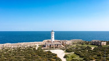 Siraküza yakınlarındaki kayalık kıyılarda çevresindeki harabeleri olan beyaz deniz feneri, derin mavi denize bakıyor ve yeşil çalılık ve patikalarla çevrelenmiş..