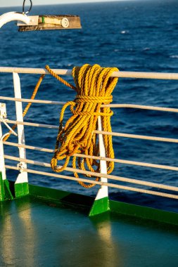 Sarmalanmış sarı halat, yeşil zeminli beyaz bir gemide asılı, sıcak ışıkla aydınlatılıyor, arka planda okyanus dalgaları görünüyor..