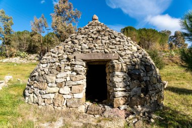 Dairesel şekilli ve küçük girişi olan kuru taş bir kulübe, parlak mavi gökyüzünün altındaki ağaçlarla çevrili çimenli bir alanda duruyor..