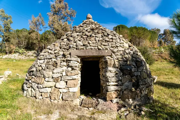 Dairesel şekilli ve küçük girişi olan kuru taş bir kulübe, parlak mavi gökyüzünün altındaki ağaçlarla çevrili çimenli bir alanda duruyor..