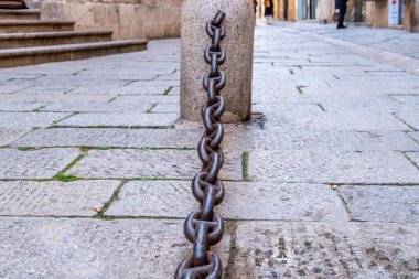 Granit bir kordonun üzerinde paslı bir zincir, düz olmayan kaldırım taşlarının üzerinde duruyor. Taş basamaklar kumtaşı bir duvara götürür, ötesinde bulanık figürler.