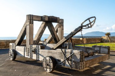 Büyük ahşap bir mancınık denize bakan asfalt bir meydanda duruyor. Metal alaşımlar ve zincirler güneş ışığıyla yapısını güçlendirir.