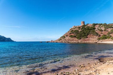 Mavi deniz, engebeli uçurumlar ve açık gökyüzünün altındaki yeşil çalılarla çevrili antik taş gözetleme kulesiyle Rocky sahil tepesi..
