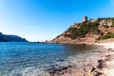 Mavi deniz, engebeli uçurumlar ve açık gökyüzünün altındaki yeşil çalılarla çevrili antik taş gözetleme kulesiyle Rocky sahil tepesi..