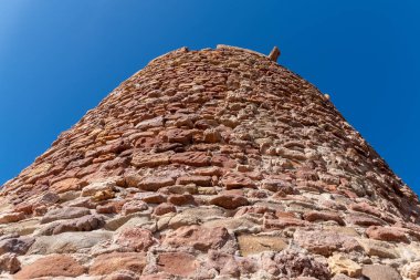 Kırmızımsı ve bej kayalardan inşa edilmiş yuvarlak taş kulenin yukarıdan görünüşü kaba dokusunu ve derin mavi bulutsuz gökyüzünün altında berrak detaylarını gösteriyor..