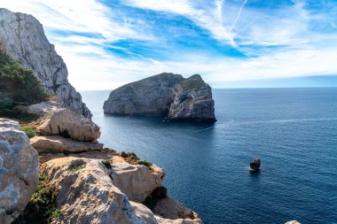 Kayalık ada derin mavi denizden yükseliyor, parlak gökyüzünün altındaki yüksek kıyı uçurumundan görünüyor dağınık bulutlar ve berrak ufuk manzarası ile.