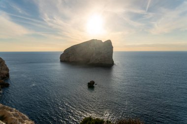 Güneşin tam üstünde olduğu sakin denizdeki büyük kaya adası, parlayan gökyüzü ve yumuşak ışık Alghero, Sardunya yakınlarındaki su yüzeyine yansıyor..