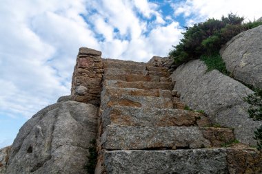 Kayalık araziye oyulmuş yıpranmış taş basamaklar kayalar ve Sardunya 'nın Palau kenti yakınlarındaki bitki örtüsü arasında yukarıya doğru parçalı bulutlu bir gökyüzünün altına çıkıyor..
