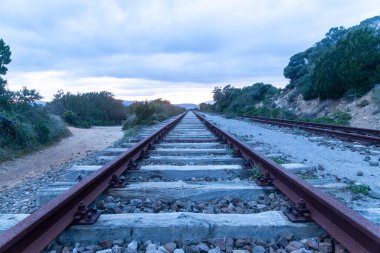 Ahşap bağlar ve çakıl döşeli paslı tren yolu, yeşil bitki örtüsü ve Golfo Aranci, Sardunya yakınlarında bulutlu gökyüzünün altında tepelerle çevrili..