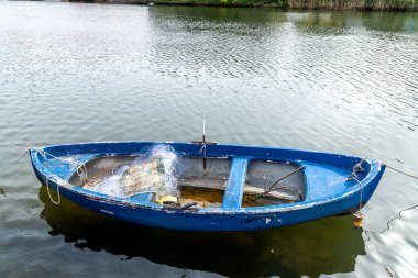 Küçük mavi bir balıkçı teknesi, görünür bir aşınmayla sakin sularda yüzer. İçinde balık ağı, halat ve gövdenin içinde tahta bir kürek bulunur..