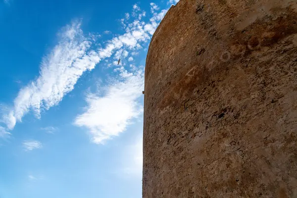 Yıpranmış yüzeyi ve solmuş yazıtları olan büyük, yuvarlak bir kireç taşı kulesi. Sağda taş yapı, bulutlu mavi gökyüzü ve solda uçan martı..