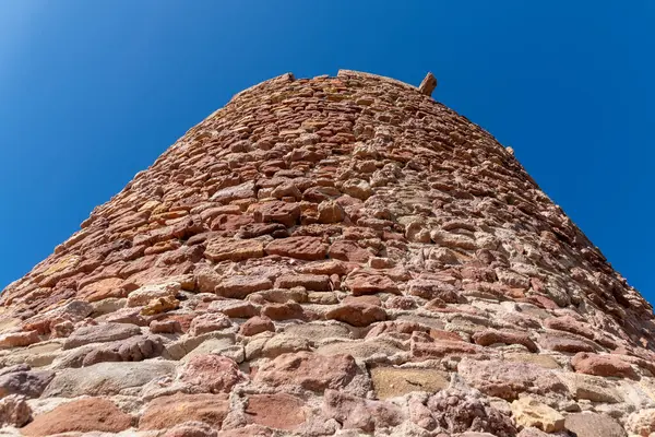 Kırmızımsı ve bej kayalardan inşa edilmiş yuvarlak taş kulenin yukarıdan görünüşü kaba dokusunu ve derin mavi bulutsuz gökyüzünün altında berrak detaylarını gösteriyor..