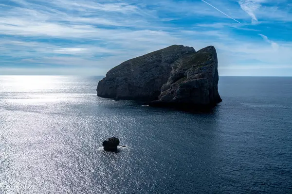 Denizdeki büyük kayalık ada parçalı bulutlu gökyüzünün altında. Etrafı sakin, pırıl pırıl suyla çevrili. Güneş ışığı yüzeyden yansıyor..