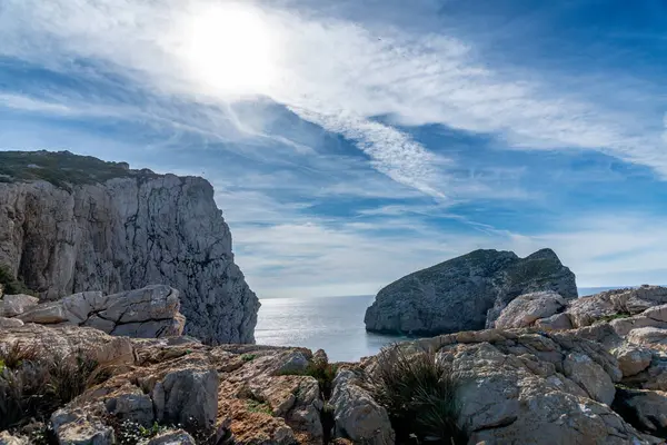 Kayalık kayalıklar ve denizden yükselen sarp bir ada parçalı bulutlu bir gökyüzünün altında güneş ışıl ışıl parlıyor..