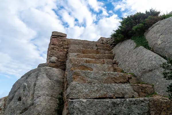 Kayalık araziye oyulmuş yıpranmış taş basamaklar kayalar ve Sardunya 'nın Palau kenti yakınlarındaki bitki örtüsü arasında yukarıya doğru parçalı bulutlu bir gökyüzünün altına çıkıyor..