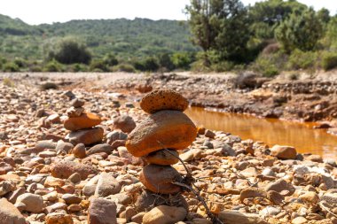 Sardinya 'daki Rio Piscinas yakınlarındaki kayalık bir nehir yatağında dengelenmiş taş yığınları, yeşil tepeler ve sığ, paslı bir dere.