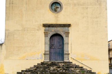 Sicilya, Sperlinga 'daki tarihi taş kilise girişi süslü ahşap bir kapısı, yıpranmış taş basamakları ve yukarıda küçük dairesel bir dini rahatlama..
