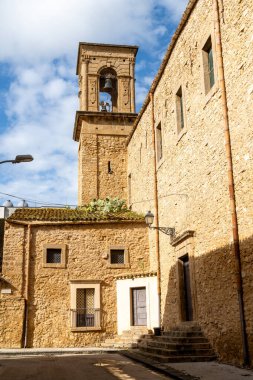 Çan kulesi ve Niscemi 'deki bitişik taş binalar mavi gökyüzünün altında, fayanslı bir çatıda kaktüsler ve eski caddeye düşen gölgeler..