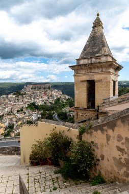 Ragusa 'nın tarihi yamaçlarına bakan çan kulesi ve taş merdiven, dağınık evler ve bulutlu bir gökyüzünün altındaki yeşil tepeler..