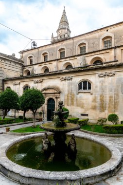 San Giovanni Battista Kilisesi 'nin dış cephesinin detaylı bir görüntüsü, ön planda bir taş çeşme ve yan tarafında süs ağaçları var..