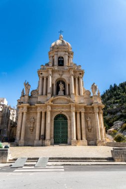 Scicli 'deki San Bartolomeo Kilisesi' nin şaşaalı cephesi. Heykeller, sütunlar ve parlak mavi gökyüzünün altında belirgin bir kubbe..