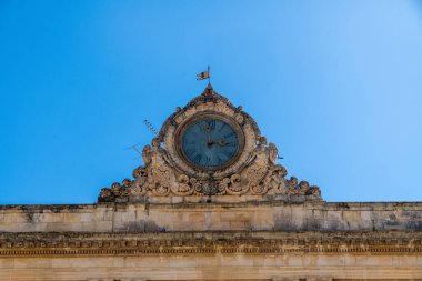 Scicli 'deki taş bina üzerindeki tarihi saati karmaşık oymalar, heykelsel detaylar, mavi gökyüzü ve klasik mimari elementlerle donatın..