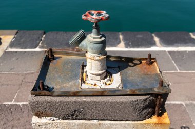 Syracuse rıhtımında beton bir bloğa monte edilmiş paslı metal su vanası. Direkt güneş ışığı altında sigara izmaritleri ve cıvatalarla çevrili..