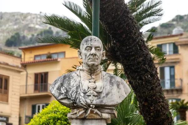 Madalyon takan bir dini figürün mermer büstü. Palmiye ağaçları ve Monreale 'nin arka planında dağlar olan binalar arasında geçiyor..