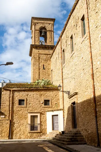 Çan kulesi ve Niscemi 'deki bitişik taş binalar mavi gökyüzünün altında, fayanslı bir çatıda kaktüsler ve eski caddeye düşen gölgeler..