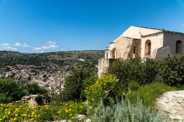 San Matteo 'dan Scicli' nin panoramik manzarası, tarihi binalar, yeşil tepeler, sarı kır çiçekleri ve arka planda parlak mavi bir gökyüzü..