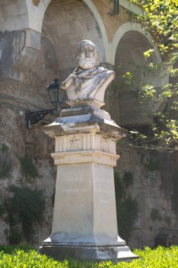 Syracuse 'da kaidede sakallı bir adamın taş büstü. Kemerli taştan bir duvarın önünde, kısmen gölgeli ve yeşilliklerle çevrili..