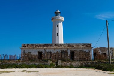 Çalılar ve mavi denizle çevrili yıpranmış taş bir yapının arkasında beyaz bir deniz feneri yükseliyor, açık gökyüzünün altında Syracuse yakınlarında çekiliyor..