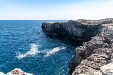 Syracuse yakınlarındaki sarp kıyı kayalıkları derin mavi denizin üzerinde dokulu kaya oluşumları ve parlak bir Akdeniz gökyüzünün altında beyaz spreyler..