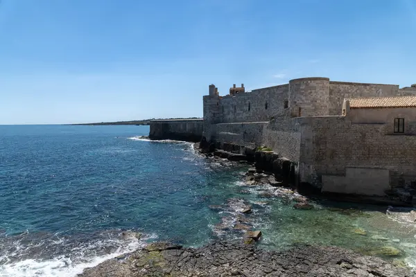 Syracuse kıyısındaki taş kale Castello Maniace, parlak mavi gökyüzünün altında turkuaz deniz ve kayalık kıyı şeridi ile çevrili..