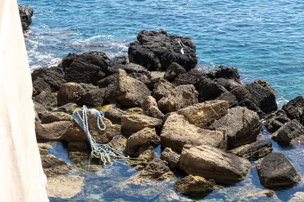 Karışık bir ip, parlak güneş ışığı altında, Syrakus kıyısı boyunca kısmen sığ sulara batmış engebeli kıyı kayaları üzerinde uzanır..