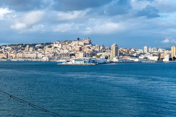 Denizi aşıp Cagliari 'nin güneşli tarihi şehir manzarasına doğru bakın. Tepenin yamacında yükselen binalar bulutlu bir gökyüzünün altında..