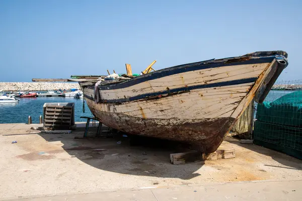 Liman bölgesinde beton destekli eski bir ahşap tekne. Tekne pas ve yıpranmayı gösteriyor, arka planda deniz, tekne ve liman duvarı var..