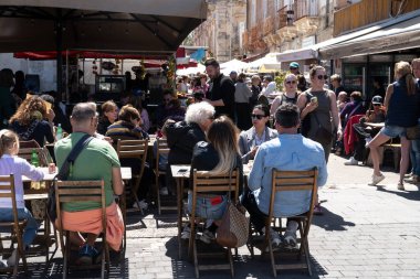 Syrakus, Sizilien - İtalya - 04-09-2025: Kalabalık Syrakus 'taki açık kafe masalarında, şemsiyeler altında ve kaldırımlı bir sokakta güneş ışığı altında konuklarla ilgilenen bir garson eşliğinde oturup yürüyor.