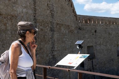 Syrakus, Sizilien - İtalya - 04-09-2025: Kadın, Syracuse 'daki tarihi taş kale yakınlarında güneş lambalı bilgilendirme tahtası okuyor, parlak güneş ışığı altında paslı korkulukların yanında duruyor.