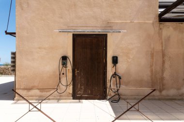 Syrakus, Sizilien - İtalya - 04-09-2025: İki elektrikli araç şarj istasyonu, Syrakus 'ta kapalı ahşap bir kapının yanında, güneş ışığının açısal gölgeleri oluşturduğu bronz bir duvara monte edildi.
