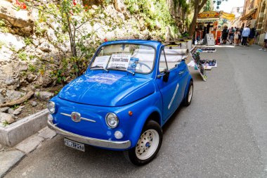 Taormina, Sizilien - İtalya - 05-04-2025: Güney İtalya 'da kayalık bir duvar ve market standının yanında yerel turlar düzenleyen yumuşak kapaklı ve ön camlı klasik mavi Fiat 500.