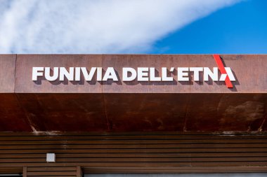 Catania, Sizilien - İtalya - 04-22-2025: Açık mavi gökyüzü altında Etna Dağı teleferik istasyonunun girişinde büyük beyaz Funivia dellEtna harfleri bulunan pas rengi çatı.