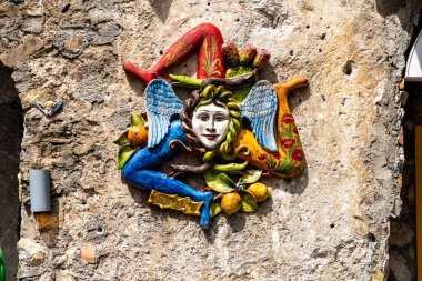 Taormina, Sizilien - İtalya - 05-04-2025: Taş bir duvara asılmış yüzlü, kanatlı, yılanlı ve bacaklı Trinacria 'nın renkli seramik dekorasyonu. Konum: Taormina, Sicilya.