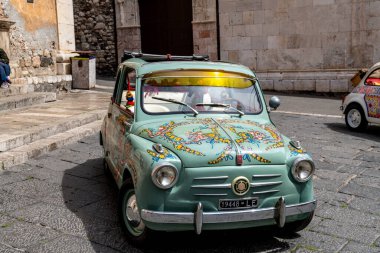Taormina, Sizilien - İtalya - 05-04-2025: Eski binaların önündeki taş kaldırıma park edilmiş Sicilya çiçekleriyle süslenmiş klasik yeşil Fiat. Konum: Taormina, Sicilya.