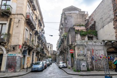 Catania, Sizilien - İtalya - 04-15-2025: Catania, Sicilya 'da dar bir sokak, eski konut binaları, balkonlar, park edilmiş arabalar, grafiti kaplı duvarlar ve şehir yeşilliği ile kaplı.